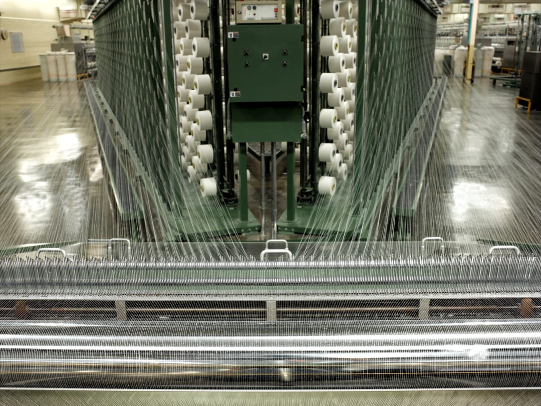 industrial weaving loom in a textile mill with rows of white cotton yarn cones feeding hundreds of warp threads