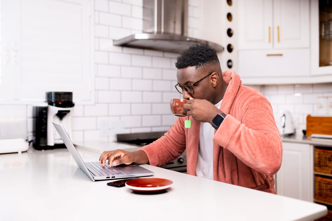 man in a soft pink plush robe drinking tea while working on a laptop at a kitchen counter