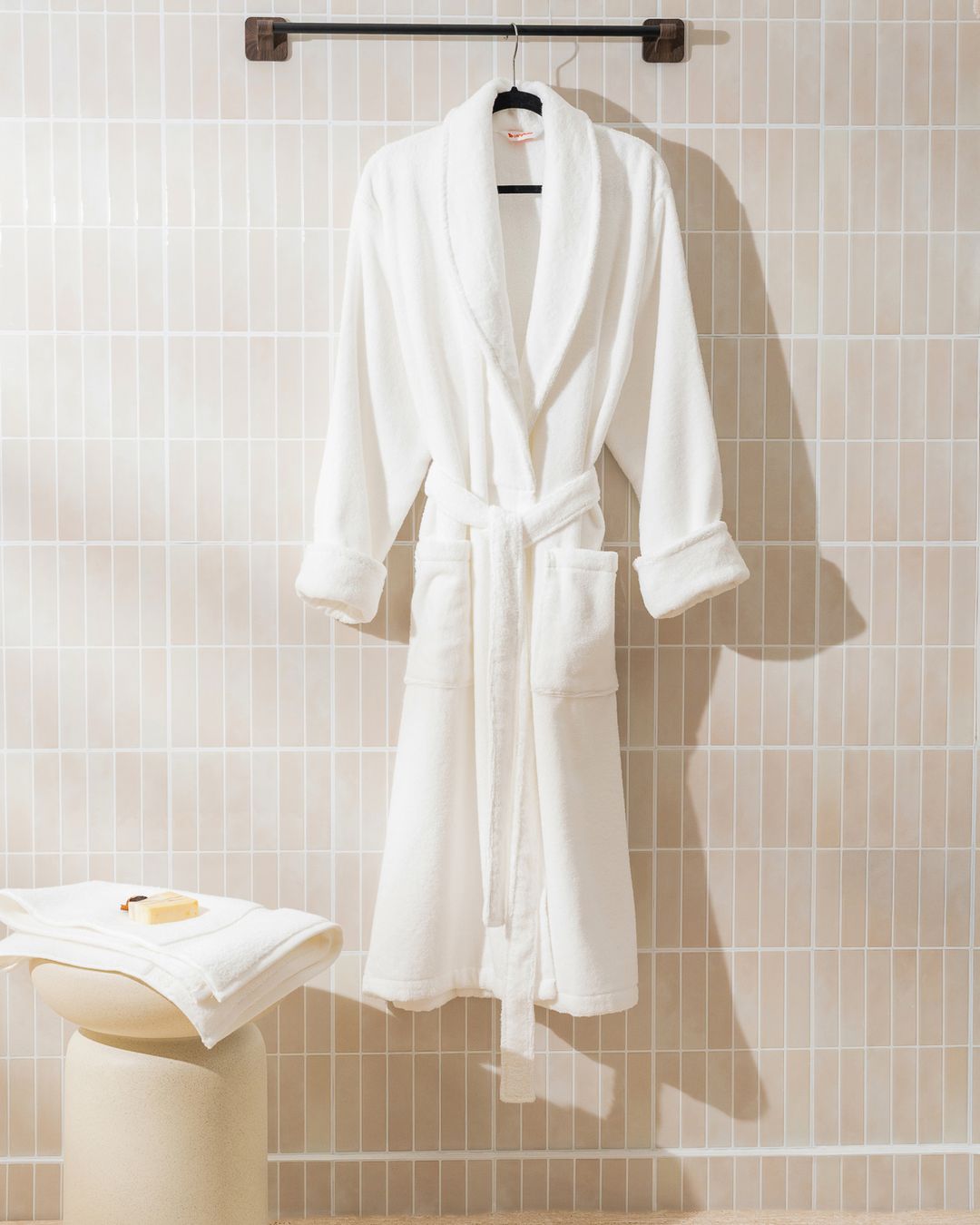 white Turkish cotton bathrobe hanging on a rail above a stool of folded towels in a tiled bathroom