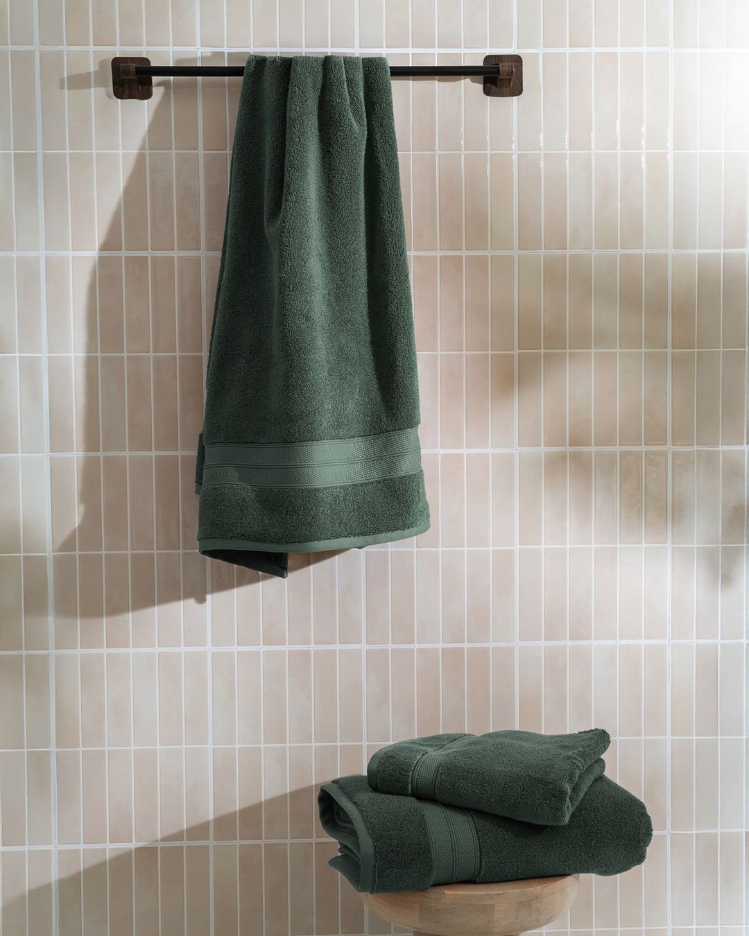 forest green modal cotton towels, one hanging on a rail and two folded on a stool in a tiled bathroom