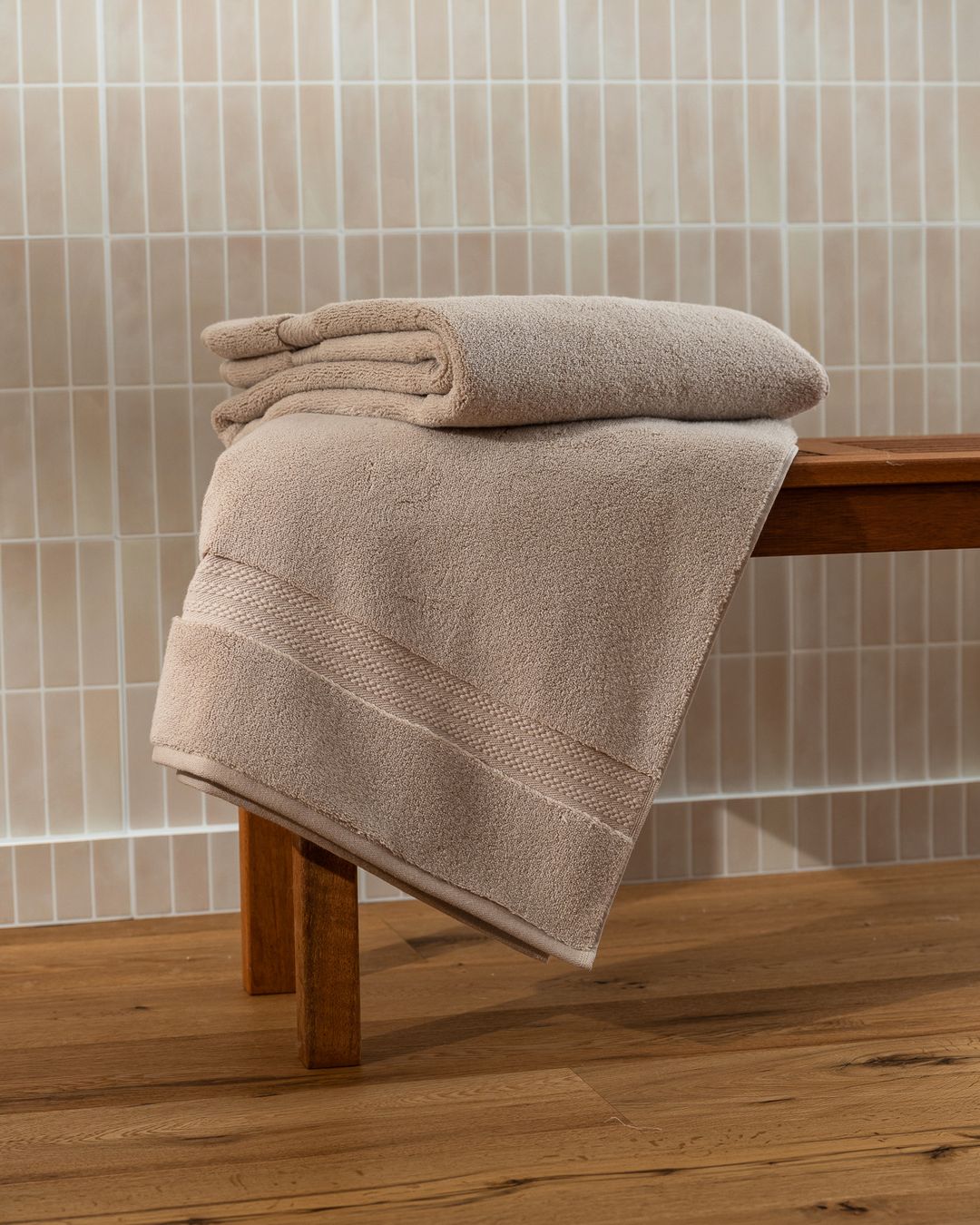 folded beige capyhome bath towel on a wooden bench in a tiled bathroom