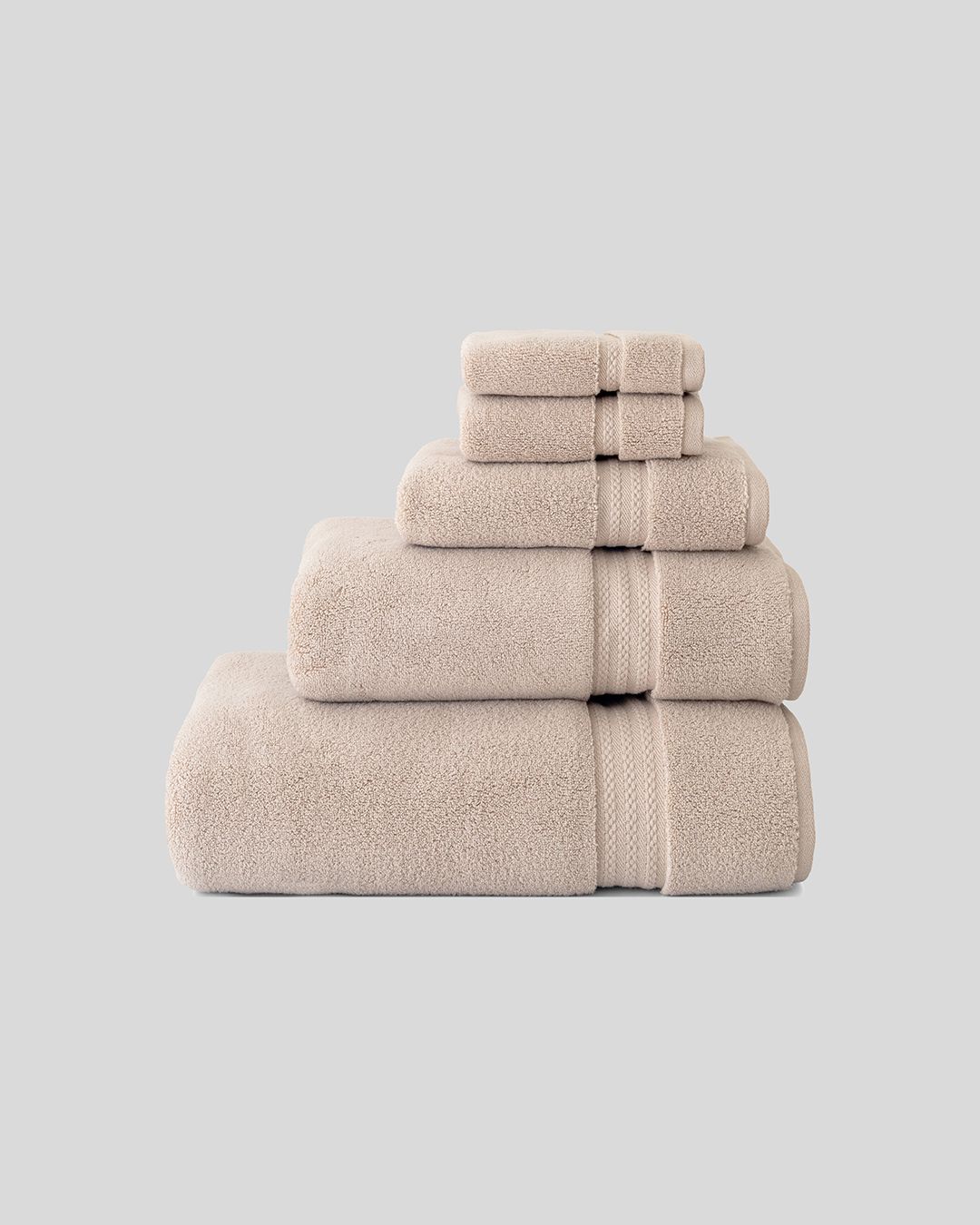 stack of folded beige terry cotton towels in five sizes, from washcloth to bath sheet, on a plain grey background